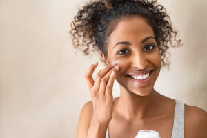 Smiling girl  applying facial moisturizer while holding jar and looking at camera. Portrait of young black woman applying cream on her face isolated on beige background. Close up of happy attractive beauty woman caring of her skin.